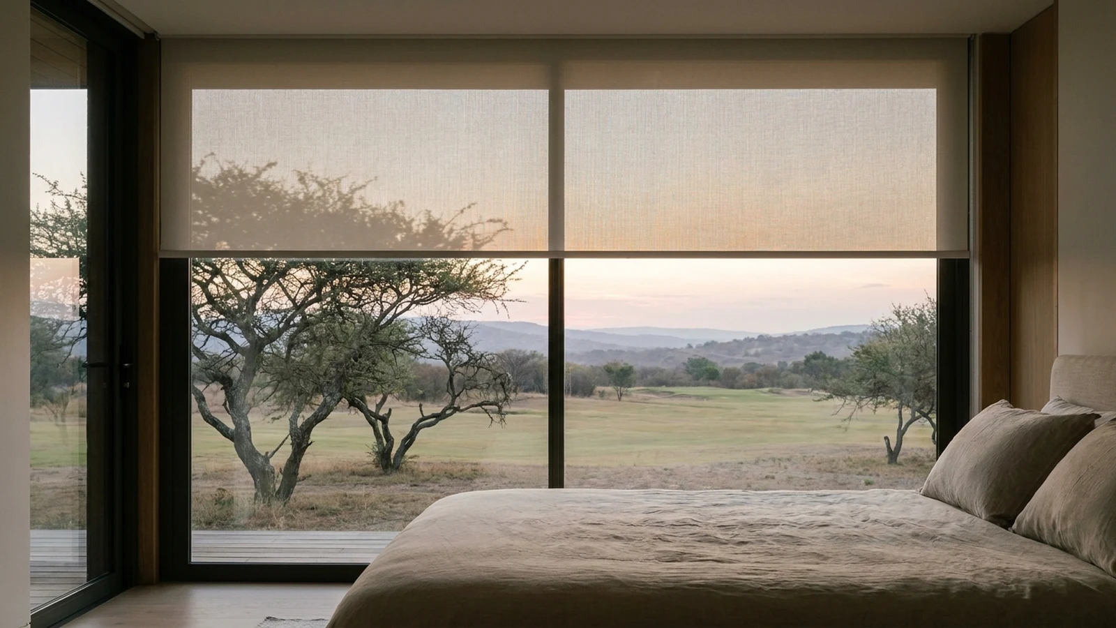 Sunscreen roller blind lowered halfway across a bedroom window at dusk, an open fairway and thorn trees fading into the distance